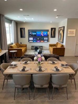 A modern living room with a flat-screen TV mounted on the wall, a dining area with a set table, and two sofas. - The Queen: 9 Bedroom/8Bath/7 En-suite Bedrooms/Sleeps 25+/LazyRiver/Pickleball (Coachella)