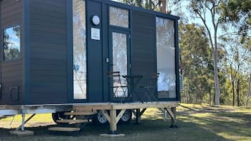 Un piccolo camper scuro con porta scorrevole, un piccolo portico con due sedie e un cartello che indica un ambiente adatto agli animali domestici.