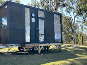 A small, dark-colored mobile home with a sliding door, a small porch area with two chairs, and a sign indicating a pet-friendly environment.