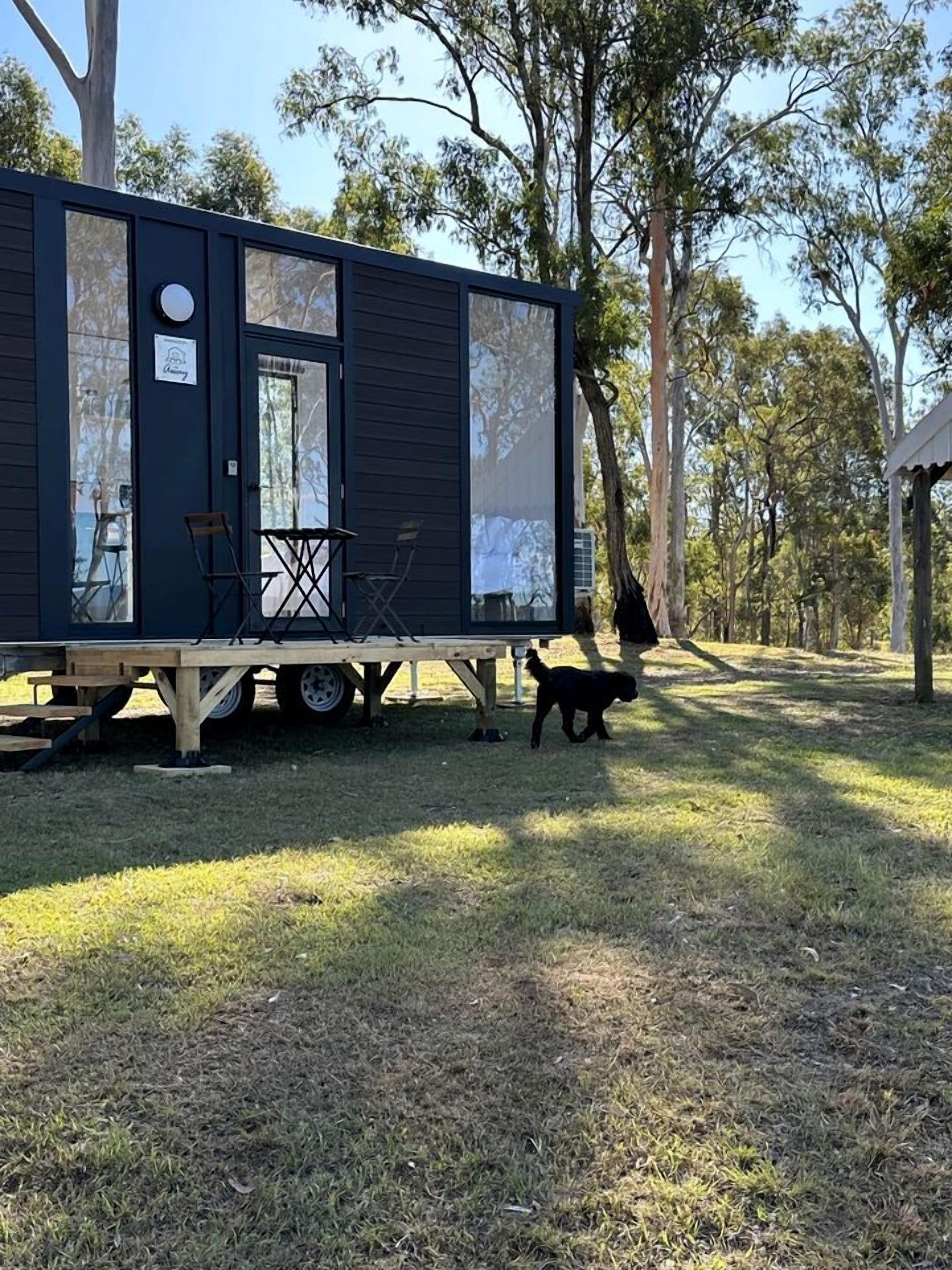 Una casa móvil moderna y de color oscuro con una pequeña terraza y un perro cerca.
