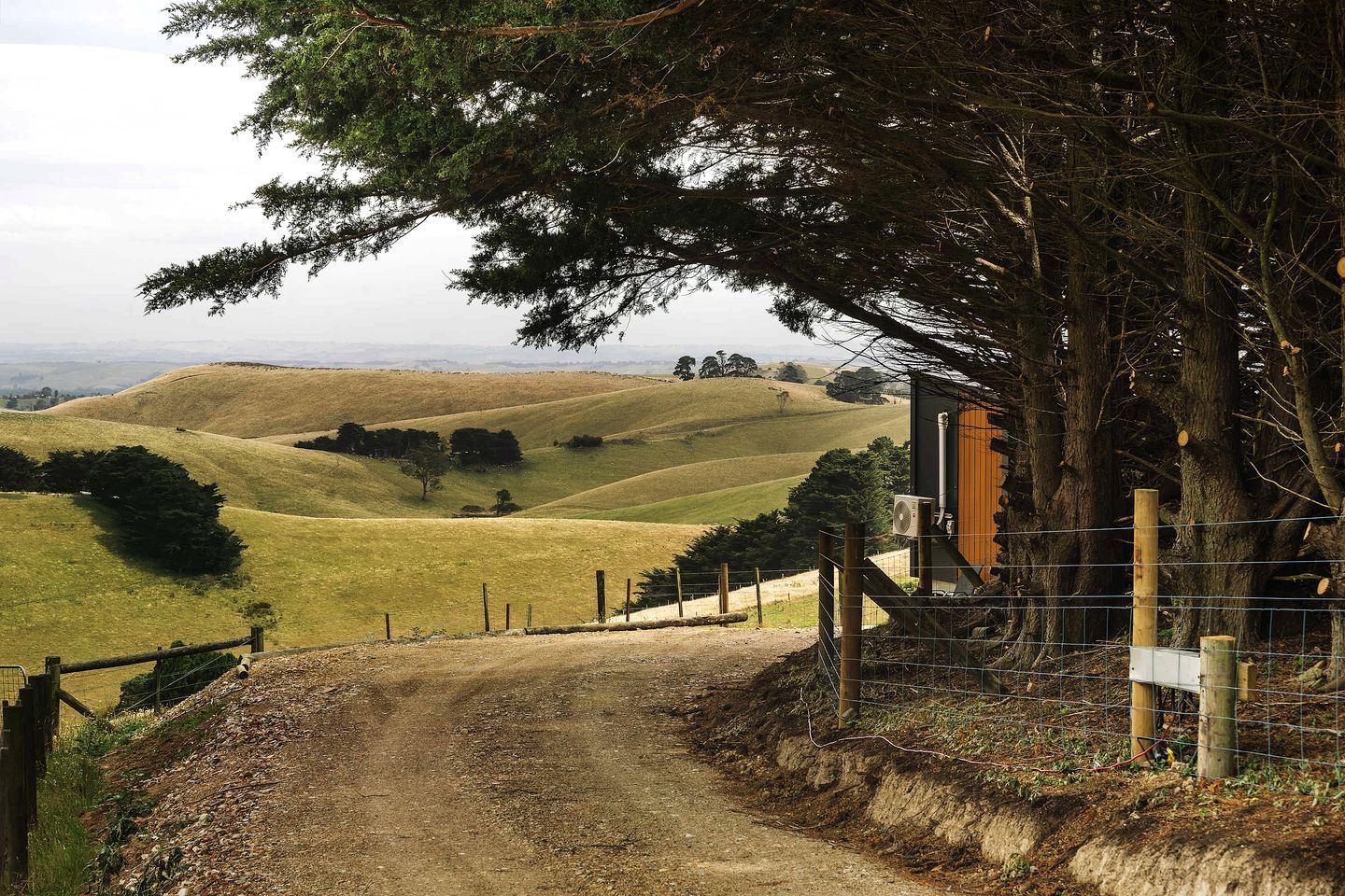 A rural landscape with rolling hills, a dirt path leading to a wooden structure, and a fenced area.