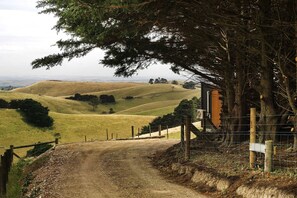 Un paisaje rural con colinas onduladas, un sendero de tierra que lleva a una construcción de madera y un área cercada.