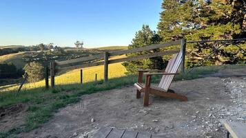 Una terraza de madera con una sola silla Adirondack, un sendero de tierra y un área cercada con árboles y un campo al fondo.