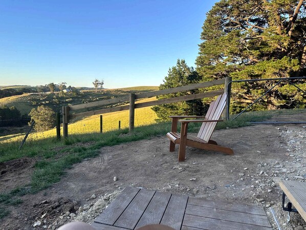 Une terrasse en bois avec un seul fauteuil Adirondack, un sentier de terre et une zone clôturée avec des arbres et un champ en arrière-plan.