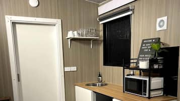 Une cuisine compacte équipée d’un four à micro-ondes, d’un évier et d’un petit plan de travail. Une porte donne accès à une autre pièce, une étagère contient des verres et un panneau d’accueil est affiché.
