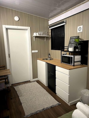 Cuina compacta amb microones, rentapelats i una petita superfície de treball. Hi ha una porta que condueix a una altra habitació, una estant amb vasos i un cartell de benvinguda.