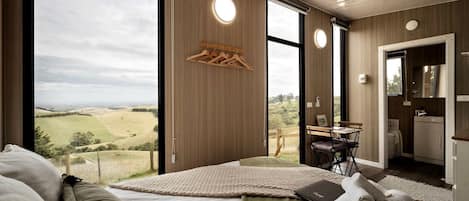 Un dormitorio moderno con un ventanal grande, una cama, una mesita y vistas al campo.