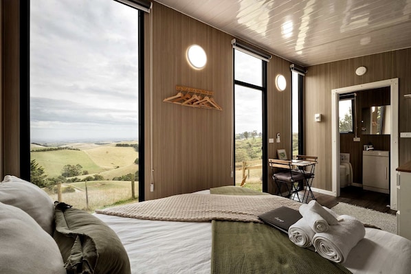 A modern bedroom with a large window, a bed, a small table, and a view of the countryside.