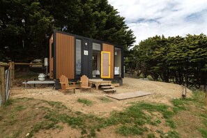 Una cabaña moderna con fachada de madera, un pequeño porche con dos sillas y una puerta amarilla.