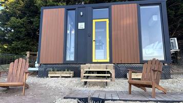 A small cabin with a yellow door, wooden walls, and a black roof. There are two wooden Adirondack chairs and a small fire pit in front.