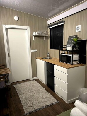 Une cuisine compacte équipée d’un four à micro-ondes, d’un évier et d’un petit plan de travail. Une porte donne accès à une autre pièce, une étagère contient des verres et un panneau d’accueil est affich é.