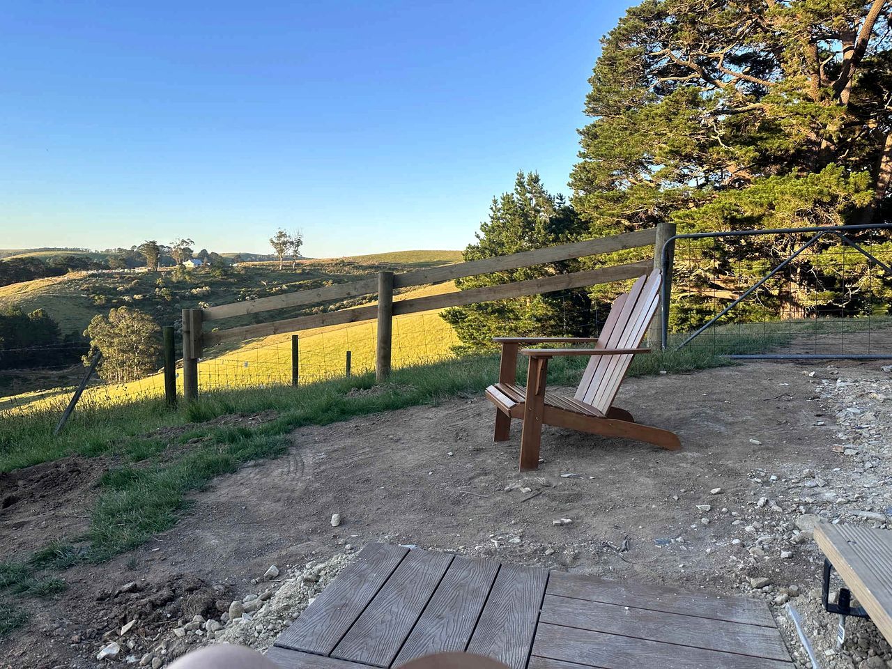 A wooden deck with a single Adirondack chair, a dirt path, and a fenced area with trees and a field in the background.