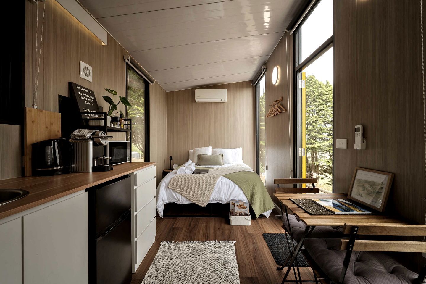 A modern bedroom with a bed, bedside table, and a view of greenery through large windows.