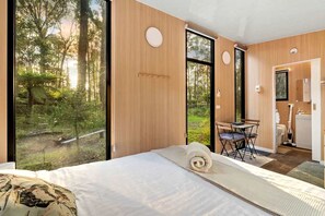 A modern bedroom with a large bed, a view of the forest, and a small desk with a chair. - Springwater Gardens Tiny Home Rental for a Yarra Valley Holiday (Toolangi)