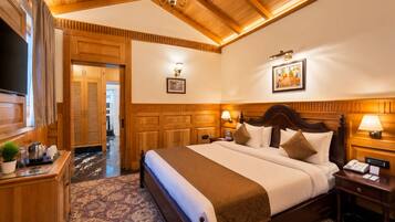 A hotel room with a large bed, wooden headboard, bedside lamps, a flat-screen TV, a wooden desk with a plant, and a patterned carpet.