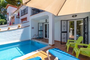 Ein Schwimmbecken mit klarer, blauer Wasseroberfläche, eine überdachte Terrasse mit gelben Stühlen und ein Gebäude mit weißer Fassade und einem Balkon.
