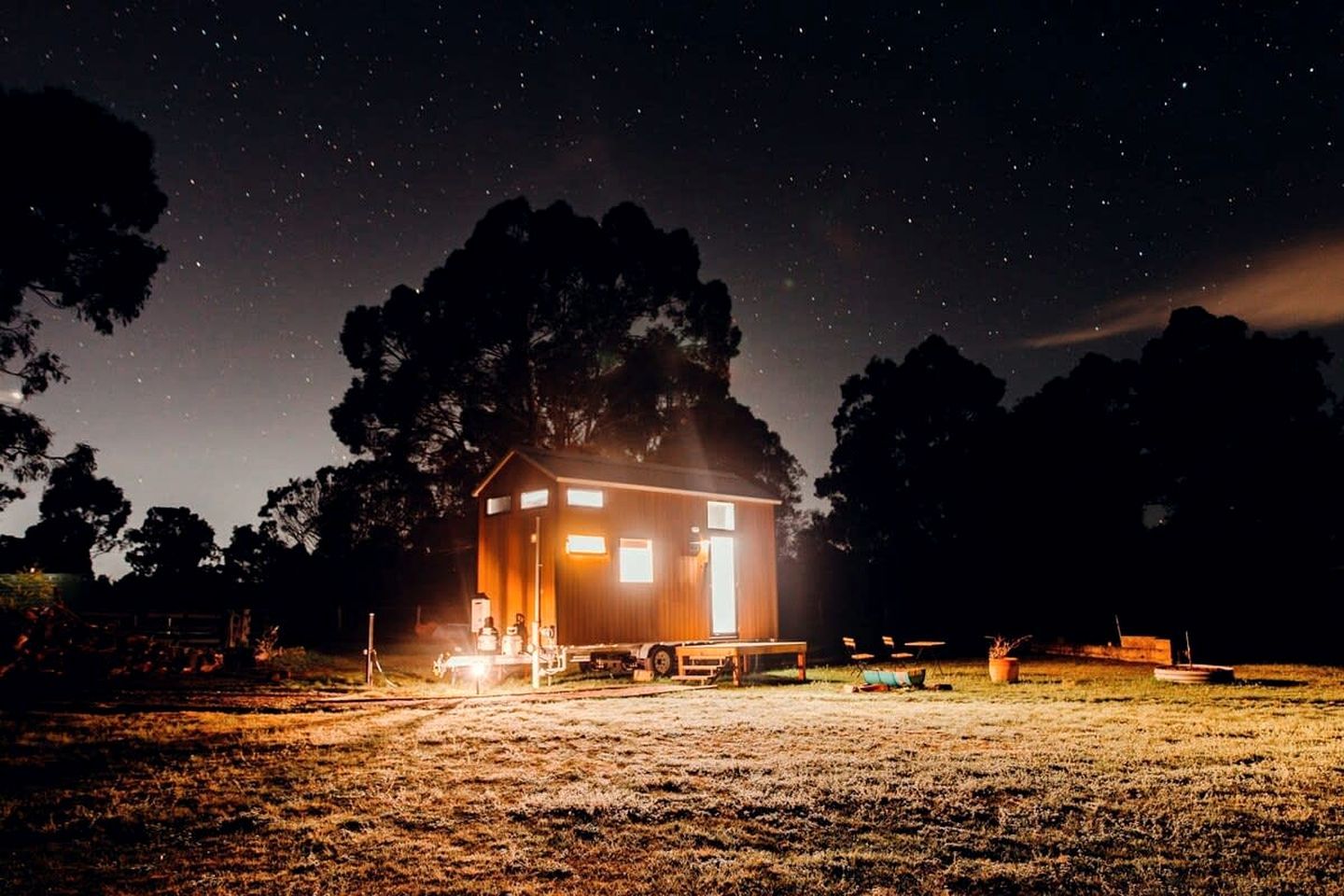 Une petite cabane éclairée, offrant une vue dégagée sur le ciel nocturne, et entourée d’arbres.