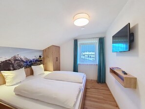 Quarto de hotel com cama, televisão fixada na parede, guarda-roupa de madeira, janela com cortinas e prateleira na parede.