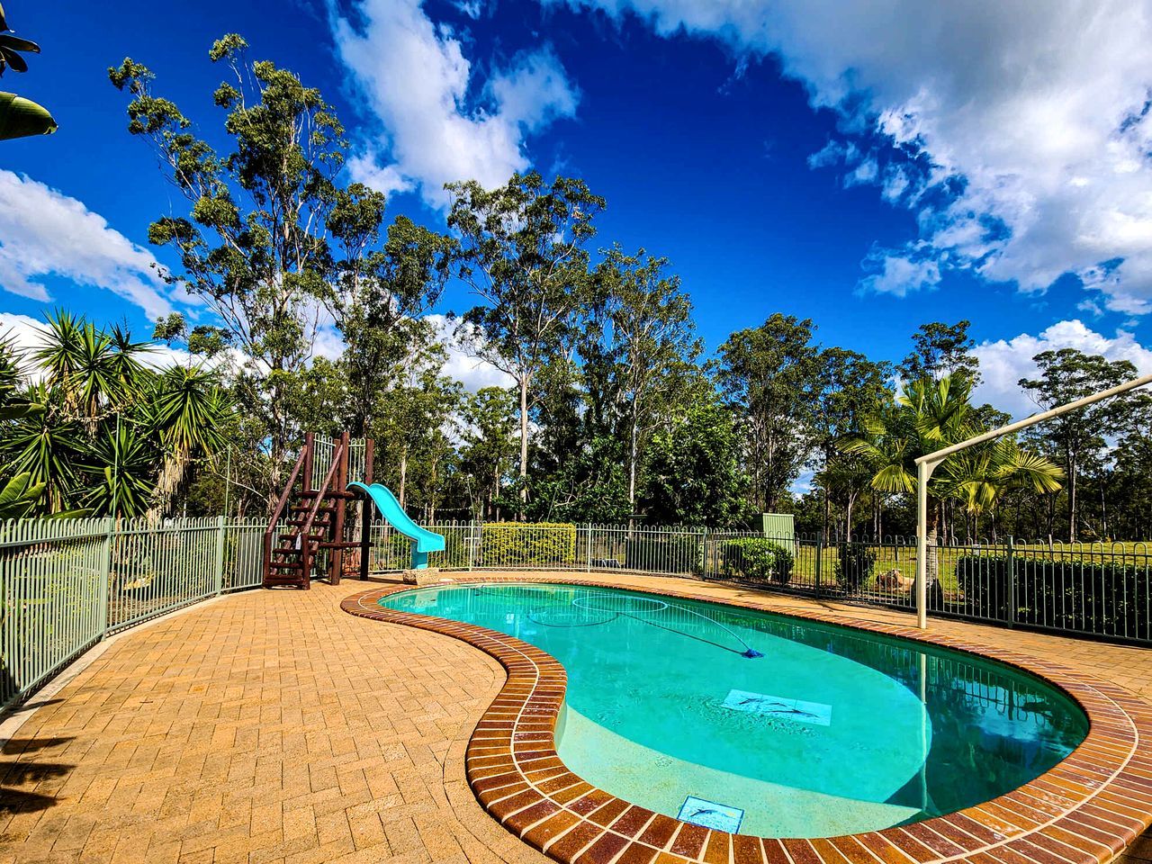 A swimming pool with a slide, surrounded by a brick walkway and a metal fence.