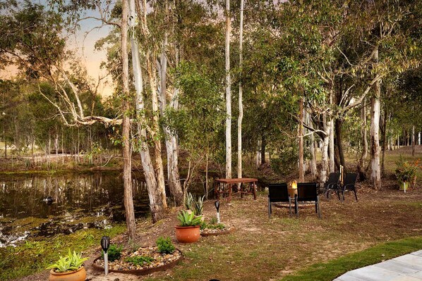 A serene outdoor setting with a wooden table and chairs, surrounded by trees and a small pond.