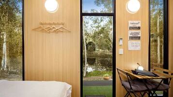 A room with a bed, a table with a chair, a wooden wall, and a view of a pond.