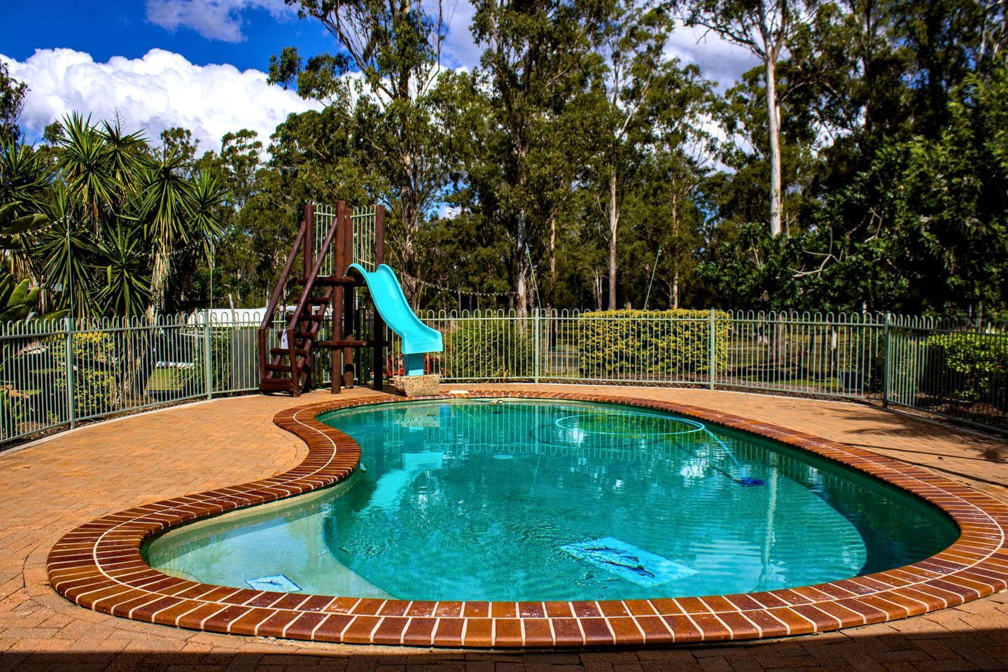 A swimming pool with a slide, surrounded by a brick border and a metal fence.