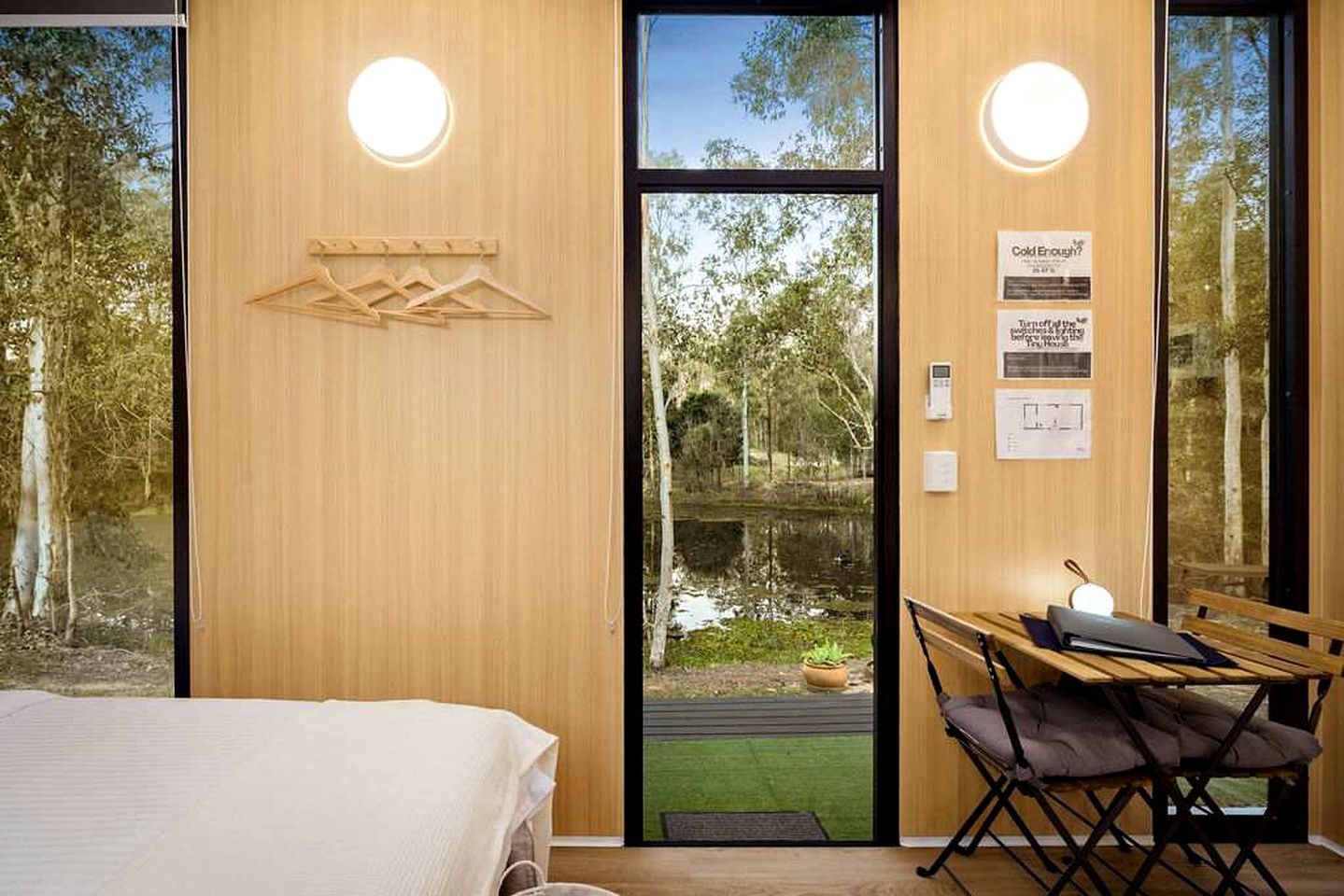 Ein Zimmer mit einem Bett, einem Tisch mit Stuhl, einer Holzwand und Blick auf einen Teich.
