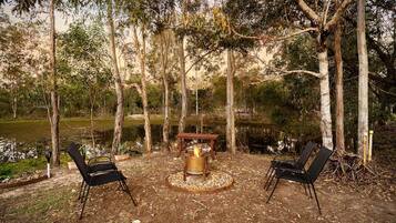 An outdoor seating area with two chairs, a table, and a fire pit surrounded by trees and a body of water.
