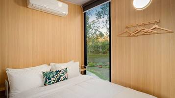A bedroom with a bed, a wall-mounted air conditioner, a window with a view of greenery, and a wooden hanger rack.