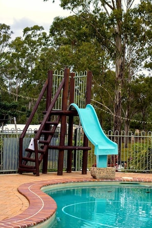 A swimming pool with a slide, surrounded by a brick deck and a metal fence.