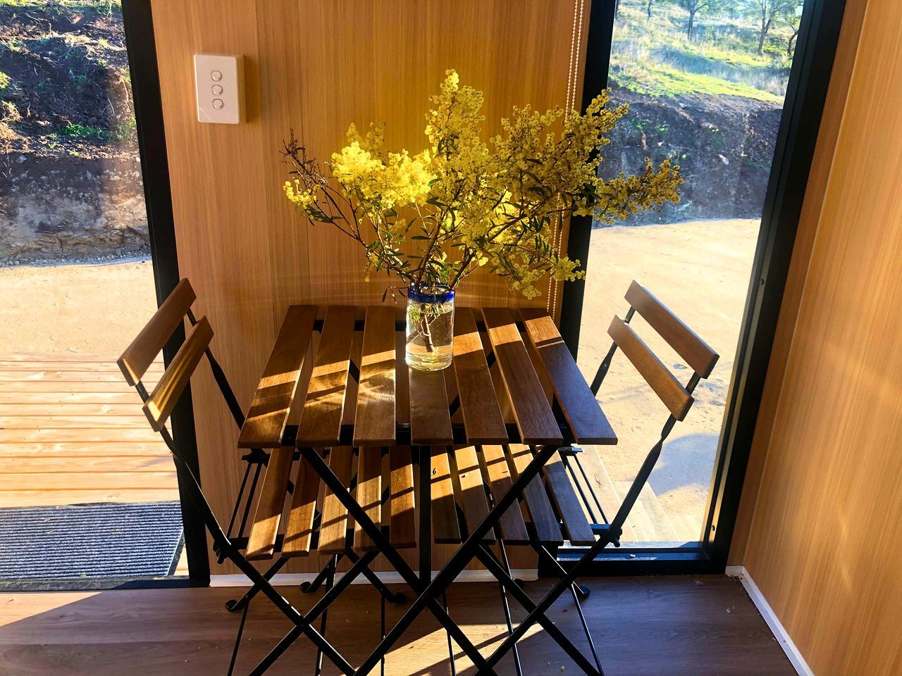 Une table en bois sur laquelle est posé un vase de fleurs jaunes, un banc et, derrière une porte vitrée, une vue sur un paysage naturel.