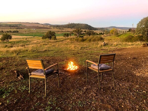 Two chairs facing a campfire in an open field with a distant view of hills and a structure.
