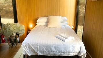 A neatly made bed with white linens and pillows, a vase with white flowers on the bedside table, and a wooden wardrobe with a hanging rack.