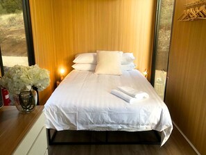 A neatly made bed with white linens and pillows, a vase with white flowers on the bedside table, and a wooden wardrobe with a hanging rack.