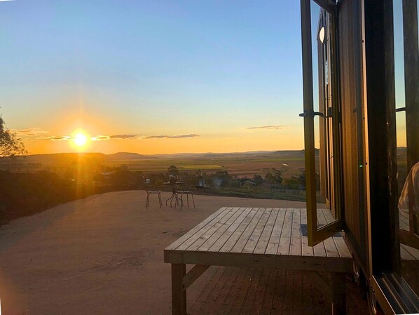 A wooden deck with a table and chairs, overlooking a landscape with fields and a sunset.