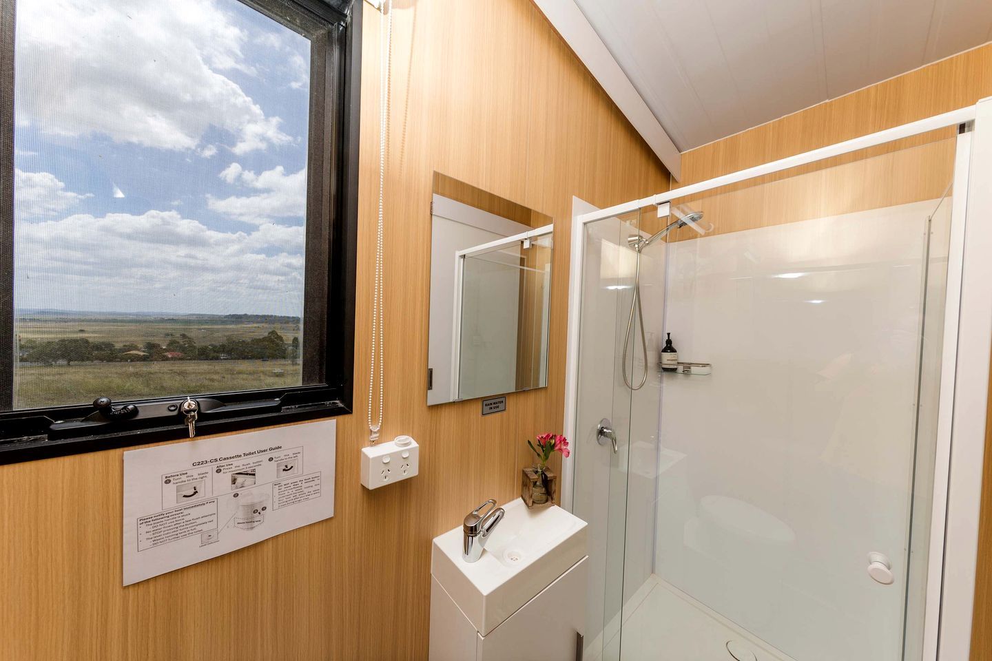 A bathroom with a shower, a sink, a mirror, and a window with a view of the countryside.