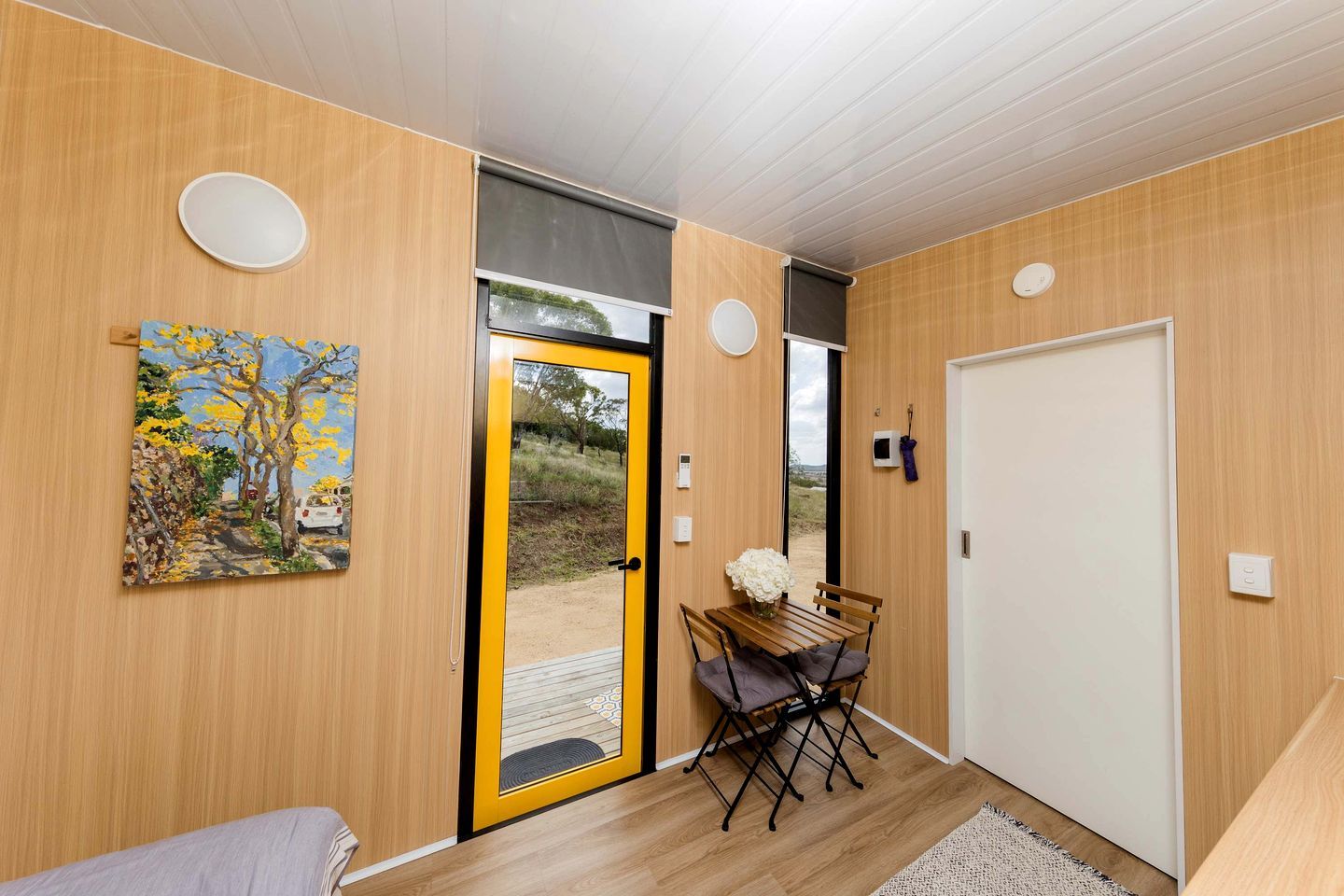 Une pièce avec un mur en bois, une porte dotée d’un cadre jaune, une petite table sur laquelle se trouve un vase, et un banc.