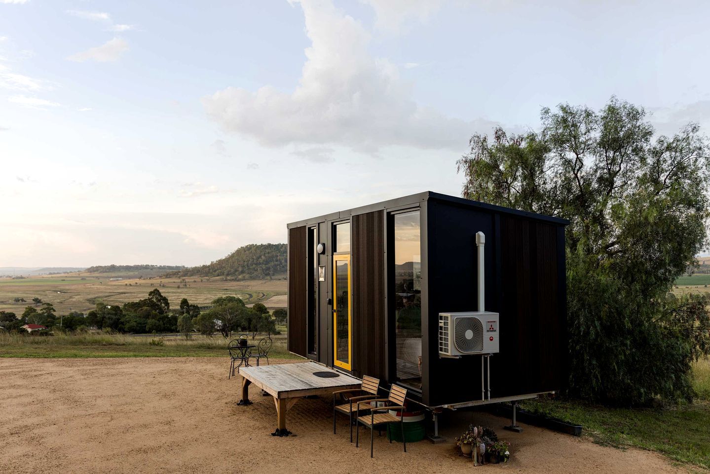 A small, dark-colored cabin with a glass door, a table, and chairs outside, set on a dirt ground with a view of a rural landscape.