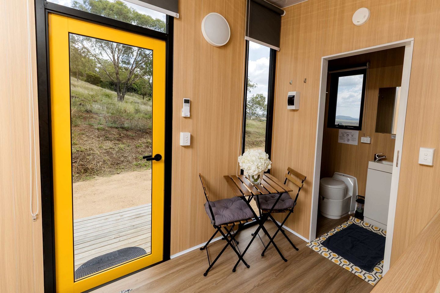 A compact room with a yellow door, a small table with a vase of flowers, a bench, a toilet, and a sink.