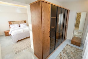 A bedroom with a wooden wardrobe, a bed with white linens, a small bedside table, and a mirror on the wall. - 2BRs Hasan Alma'moon Fabulous Apartment (Cairo Governorate)