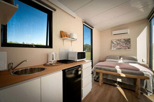A compact room with a kitchenette, a bed with a wooden frame, a window with a view of greenery, and a wall-mounted air conditioner. - Relaxing Tiny House Perfect for Horse Lovers with Firepit in New South Wales, Australia (Glenreagh)