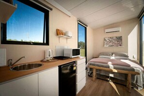 A compact room with a kitchenette, a bed with a wooden frame, a window with a view of greenery, and a wall-mounted air conditioner.