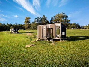 A small, single-story structure with a door and windows, situated in a grassy field with trees in the background.