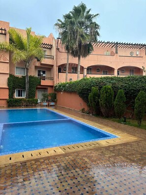 A swimming pool with a tiled deck, surrounded by a building with multiple balconies and a palm tree. - Immiouadar Elegant apartment piscine, 2min/beach (AGADIR)