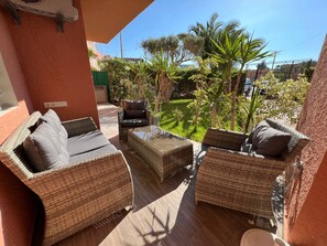 A patio with wicker furniture, a glass table, and a view of a garden with palm trees. - Immiouadar Elegant apartment piscine, 2min/beach (AGADIR)
