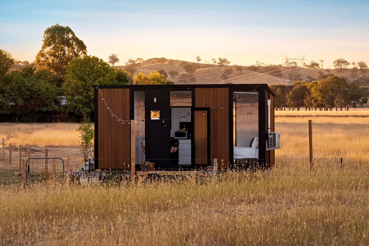 A small cabin with a kitchen area, a sleeping area, and a view of a grassy field and trees in the background.