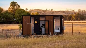 A small cabin with a kitchen area, a sleeping area, and a view of a grassy field and trees in the background.