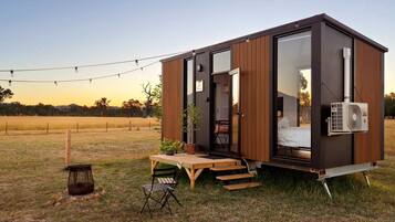 A modern, compact cabin with a large sliding door, a small porch with a table and chairs, and an air conditioning unit mounted on the exterior wall.