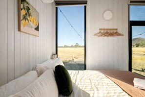 A bedroom with a bed, a window with a view of a field, and a painting of lemons on the wall. - Cozy Tiny House with Firepit Under Starry Skies in Maindample, Australia (Maindample)