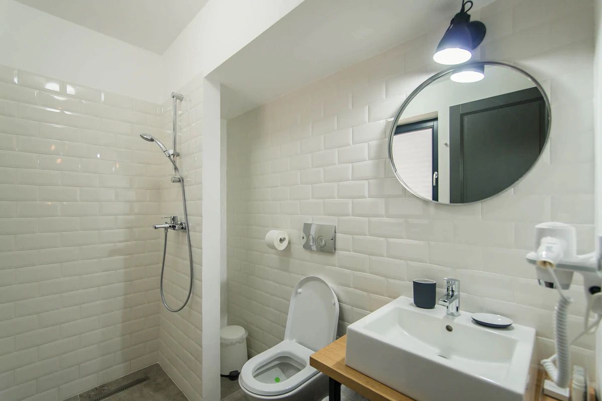 A bathroom with white tiled walls, a shower, a toilet, and a sink with a mirror above it.